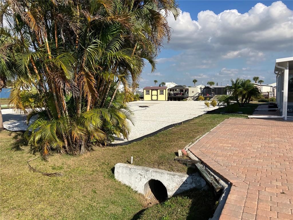A gravel yard features a small yellow shed, palm trees, and a concrete drainage culvert — 1447 FLAMINGO DRIVE, ENGLEWOOD, FL