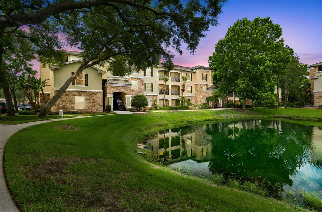 A multi-story apartment building sits behind a pond with a grassy lawn and sidewalk — 10203 COURTNEY PALMS BOULEVARD UNIT 103, TAMPA, FL