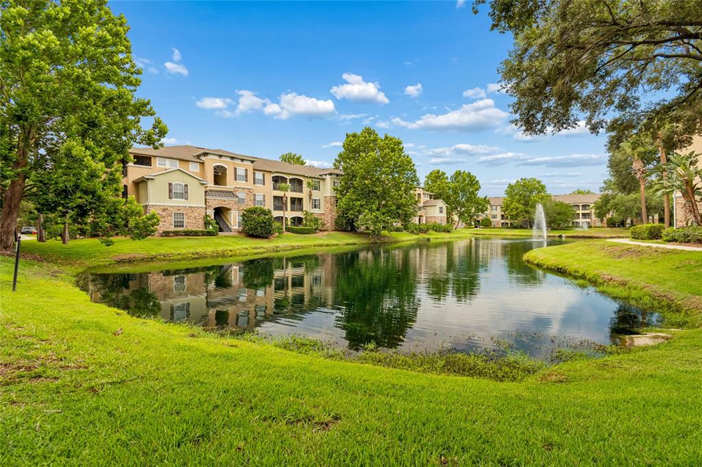 A multi-story apartment building sits behind a pond with a fountain and grassy lawn — 10203 COURTNEY PALMS BOULEVARD UNIT 103, TAMPA, FL