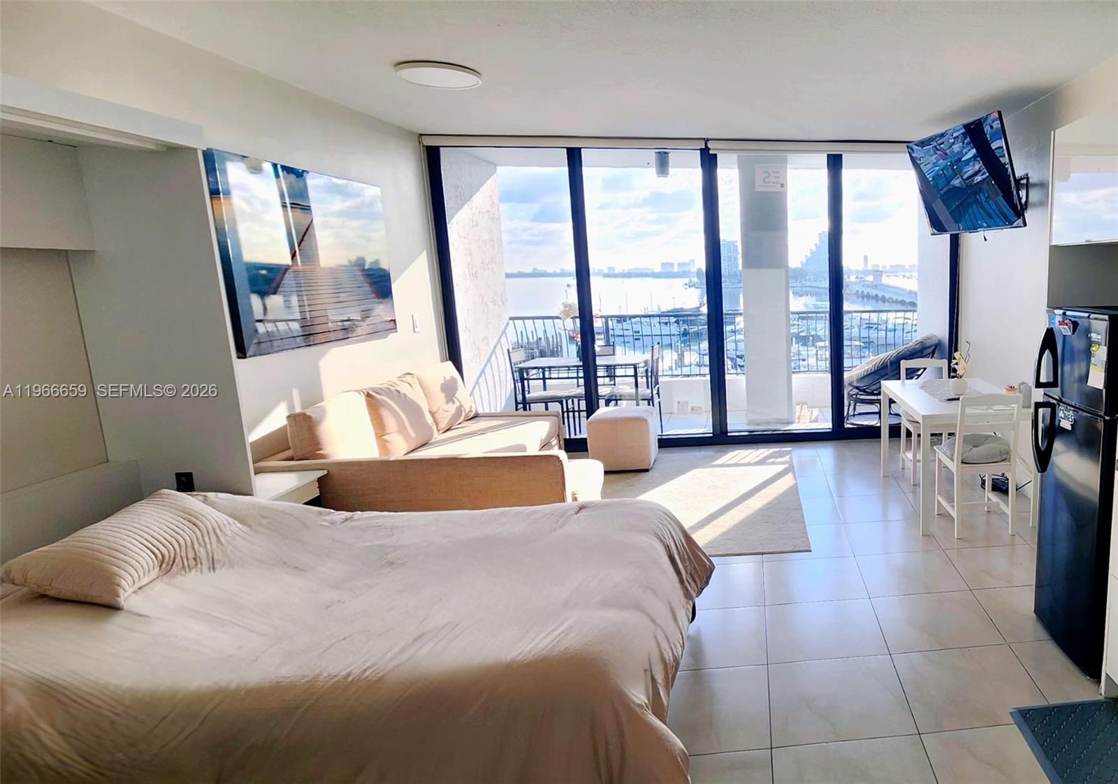 A studio apartment features a bed, sofa, dining area, and floor-to-ceiling windows overlooking water — 555 NE 15th St # 516, Miami FL 33132, MIAMI, FL