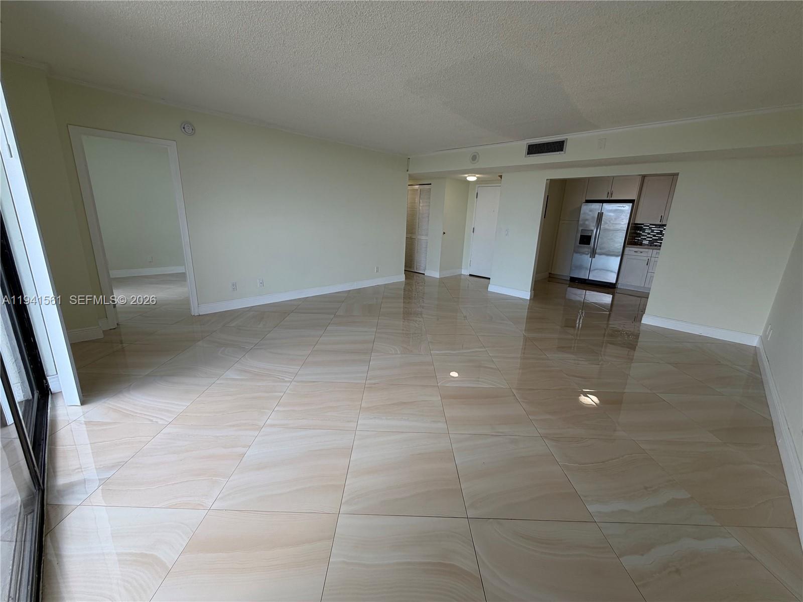 An empty living area features large beige tile flooring and an adjacent kitchen — 13499 Biscayne Blvd # 710, North Miami FL 33181, NORTH MIAMI, FL