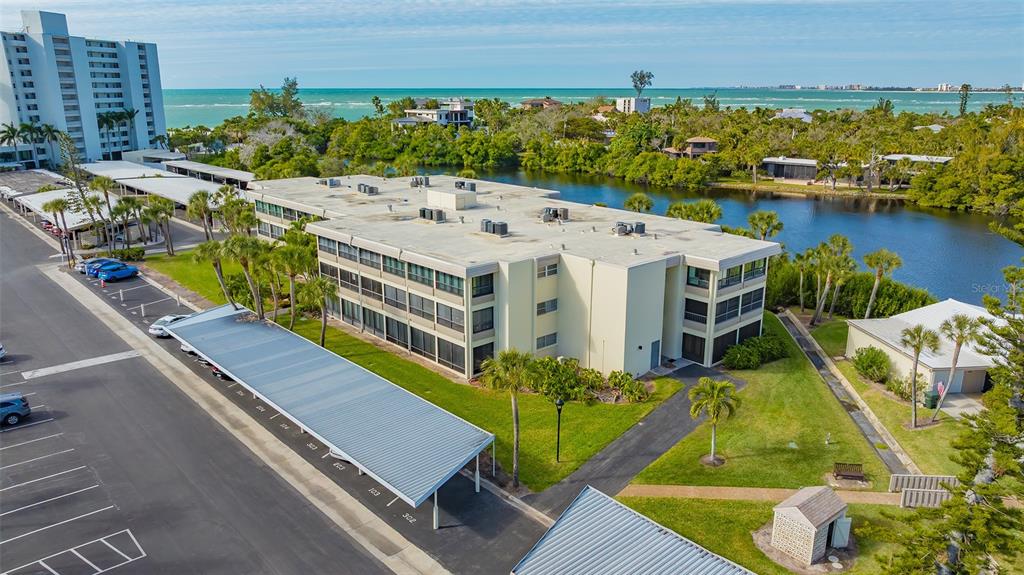 An aerial view shows a multi-story apartment building with a parking lot and water views — 101 WHISPERING SANDS DRIVE UNIT 107, SARASOTA, FL