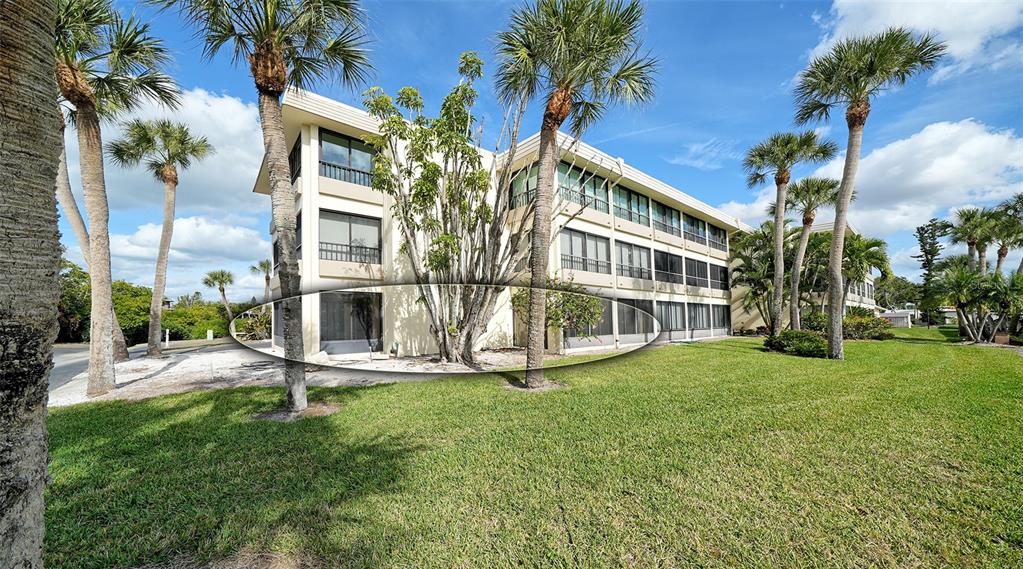 The exterior of a multi-story apartment building features palm trees and a large central tree — 101 WHISPERING SANDS DRIVE UNIT 107, SARASOTA, FL