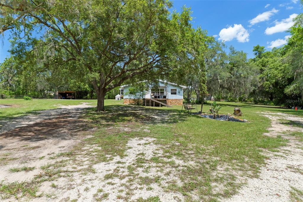 A light blue house sits on a grassy lot shaded by large oak trees — 12042 KAMBACK DRIVE, BROOKSVILLE, FL