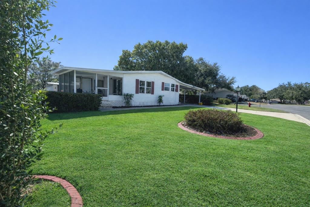 A single-story mobile home with a screened-in porch, carport, and a landscaped front lawn — 25308 CASA DEL LAGO LANE, LEESBURG, FL