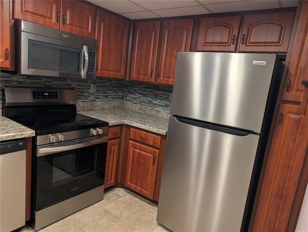 A kitchen features wood cabinets, stainless steel appliances, a tiled backsplash, and granite countertops — 3006 CARING WAY UNIT 209, PORT CHARLOTTE, FL