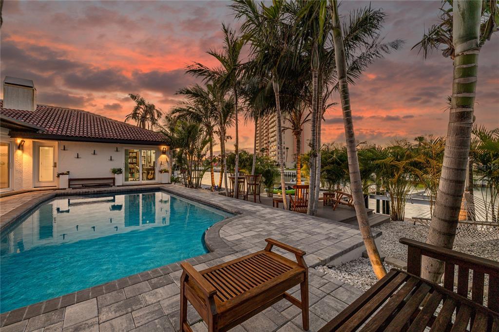 A swimming pool sits on a paved patio beside a house with palm trees at sunset — 4959 58TH AVENUE S, ST PETERSBURG, FL