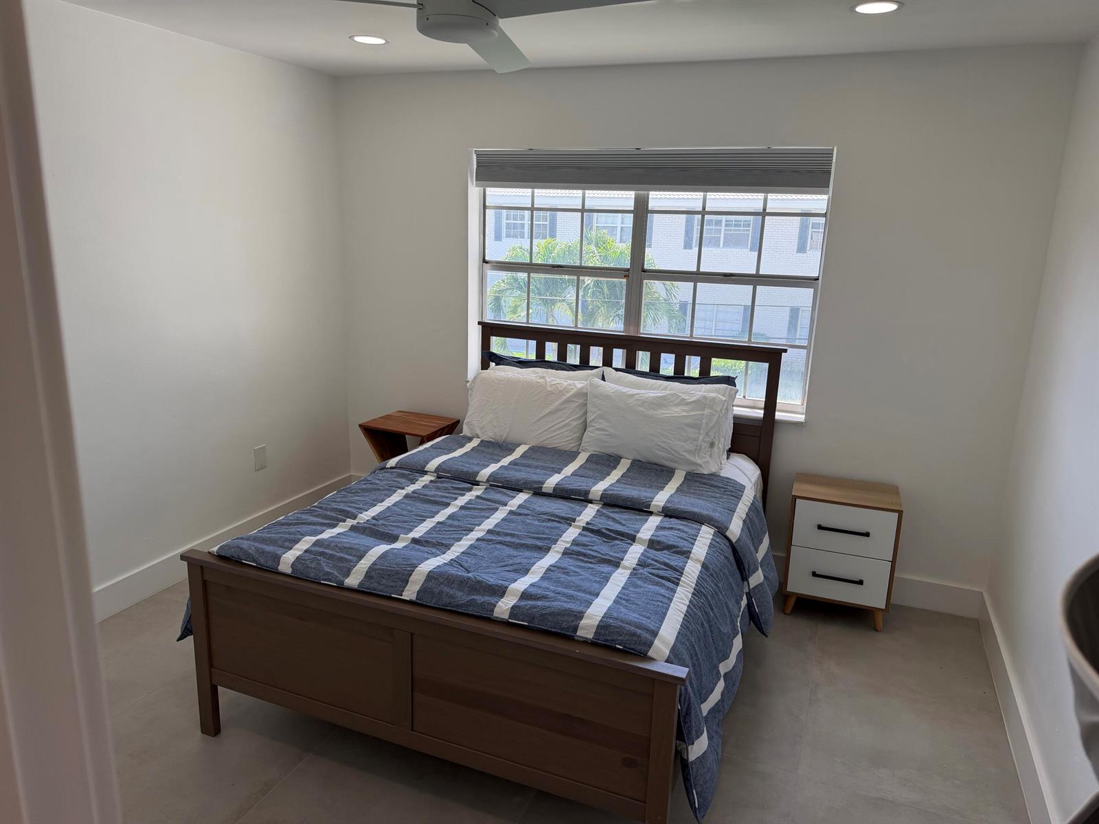A bedroom with a wooden bed, striped bedding, two mismatched nightstands, and a large window — 298 SW 6th Street # 2080, Boca Raton FL 33432, BOCA RATON, FL