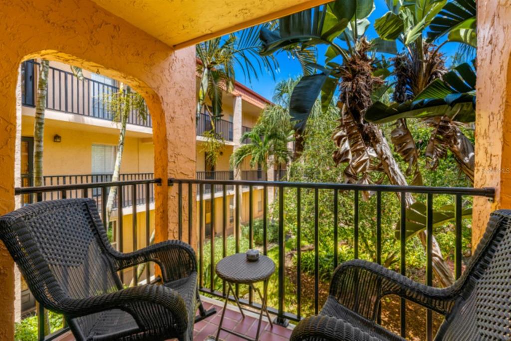A balcony with two wicker chairs and a small table overlooks a lush courtyard — 8911 BLIND PASS ROAD UNIT 223, ST PETE BEACH, FL