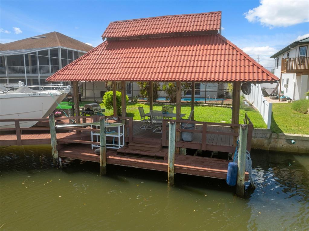 A wooden boat dock with a red-tiled roof sits over a canal near a house — 7711 HATTERAS DRIVE, HUDSON, FL