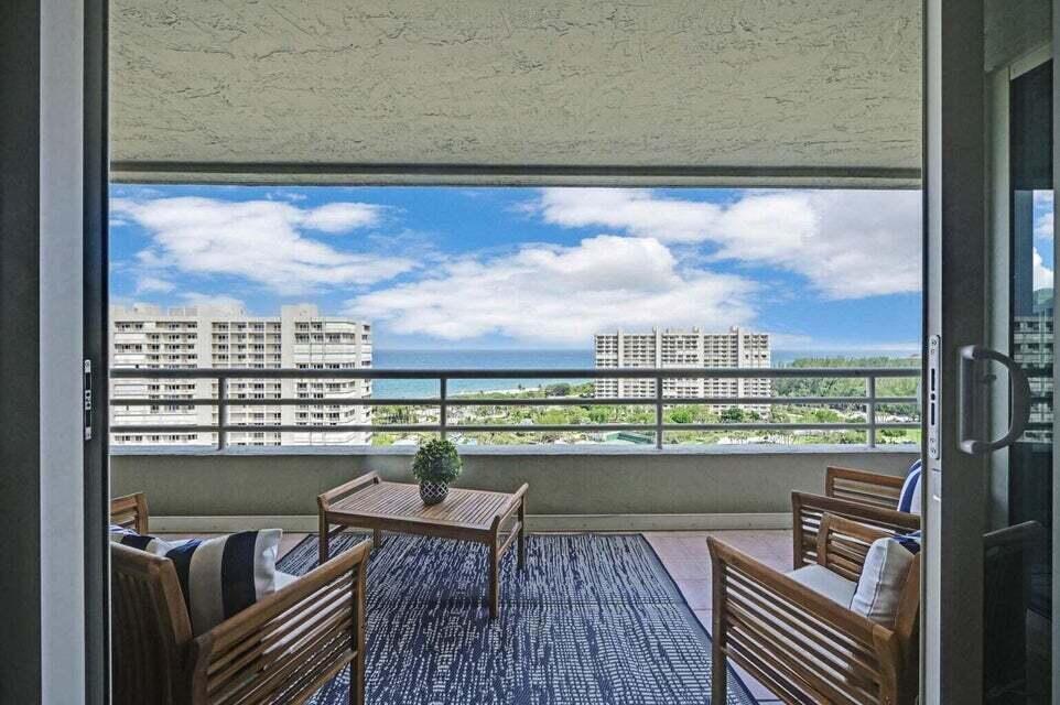 A balcony with wooden furniture and a rug overlooks the ocean and nearby high-rise buildings — 4201 N Ocean Boulevard # 1605, Boca Raton FL 33431, BOCA RATON, FL