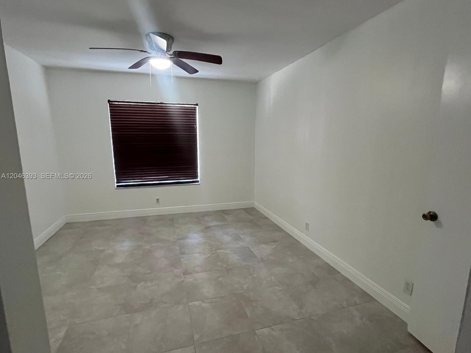 An empty bedroom features white walls, light-colored tile flooring, a ceiling fan, and a window — MIAMI, FL