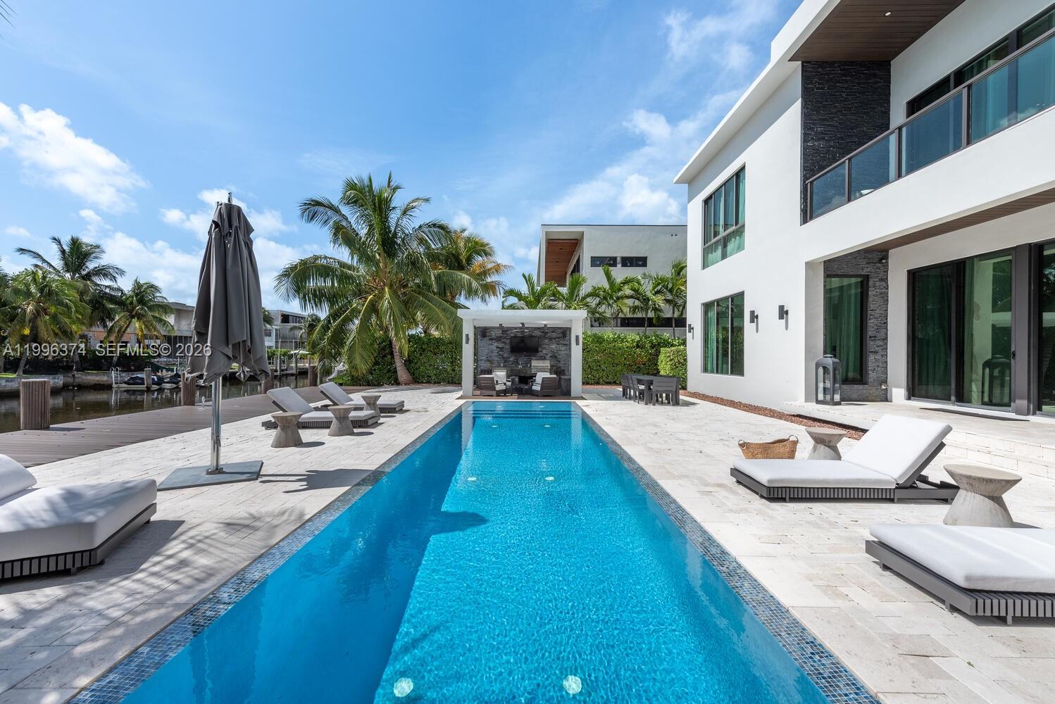 A modern house features a long rectangular swimming pool, patio furniture, and a waterfront deck — 13145 Coronado Ln, North Miami FL 33181, NORTH MIAMI, FL