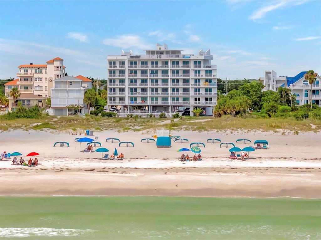 A multi-story white condominium building sits directly behind a sandy beach with beach umbrellas — 10116 GULF BOULEVARD UNIT 502W, TREASURE ISLAND, FL