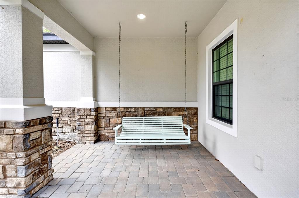 A covered porch features a light-colored hanging bench, stone accents, and paved flooring — 2612 FRAZEEK COURT, CLERMONT, FL