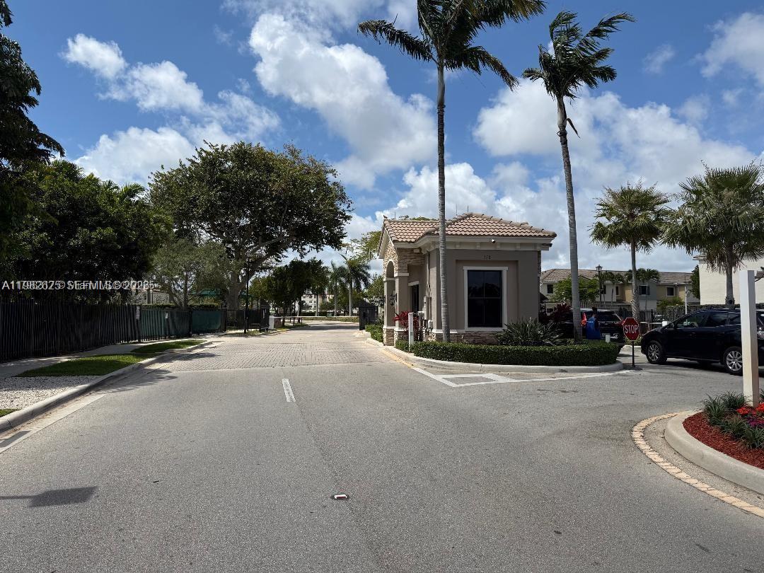 A gated entrance with a guardhouse, palm trees, and a paved road under a blue sky — 424 NE 194th Ter, Miami FL 33179, MIAMI, FL
