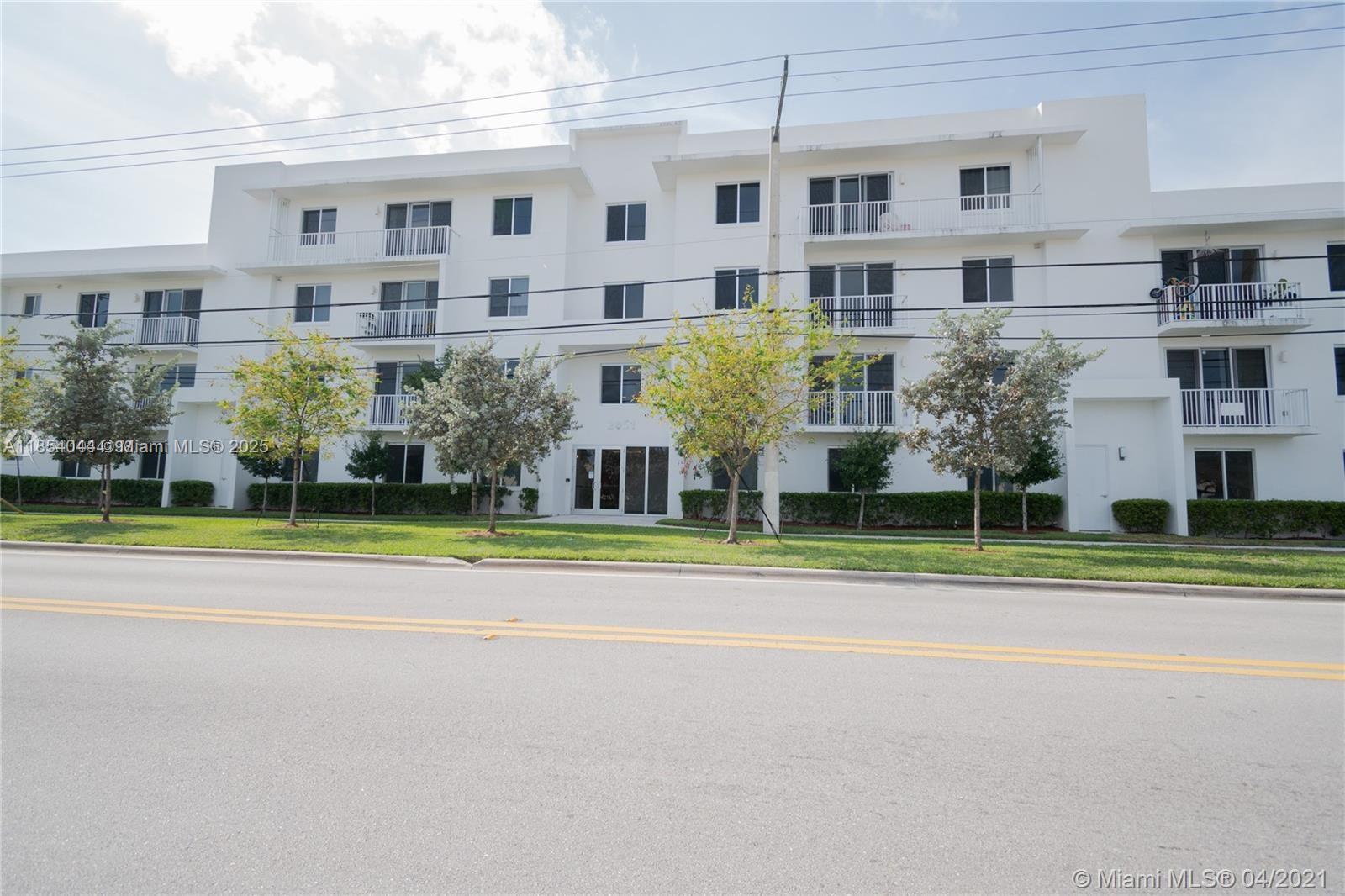 A three-story white apartment building with balconies stands behind a grassy lawn and street — 2651 NE 212th Ter # 103, Miami FL 33180, MIAMI, FL