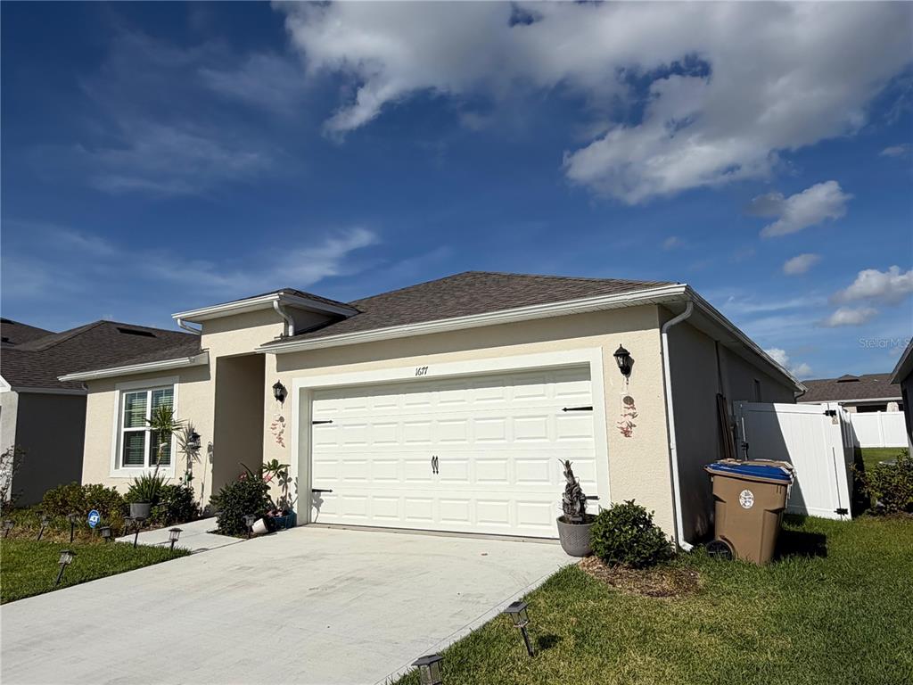 A single-story house exterior features a two-car garage, beige stucco walls, and a concrete driveway — 1677 THREE BARS ROAD, KISSIMMEE, FL