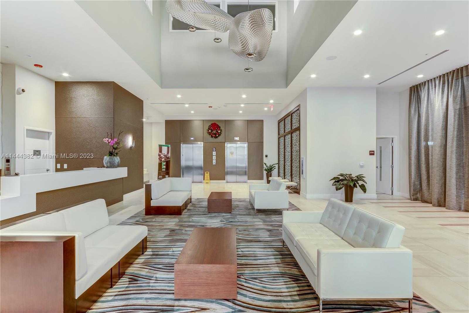 A modern building lobby features white sofas, wood coffee tables, elevators, and a large sculptural light — 7825 NW 107th Ave # 816, Doral FL 33178, DORAL, FL