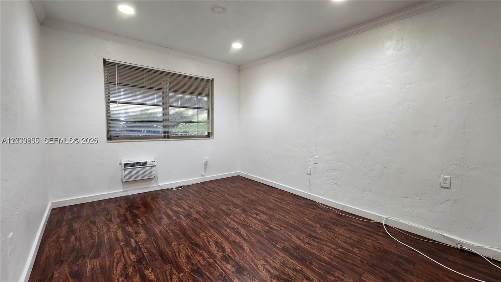 An empty room features dark wood-look flooring, white walls, a window with blinds, and an AC unit — 20 Park Dr # 4A, Bal Harbour FL 33154, BAL HARBOUR, FL