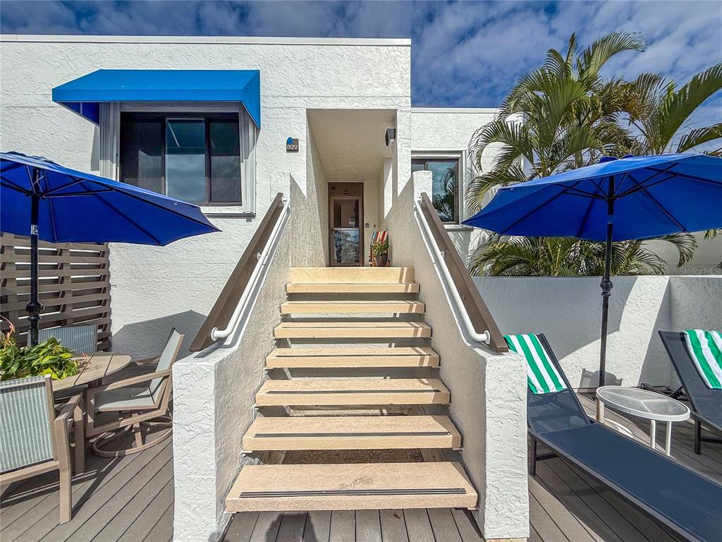 Exterior stairs lead to an entrance between two patios with blue umbrellas and lounge chairs — 829 BAYPORT WAY UNIT 829, LONGBOAT KEY, FL
