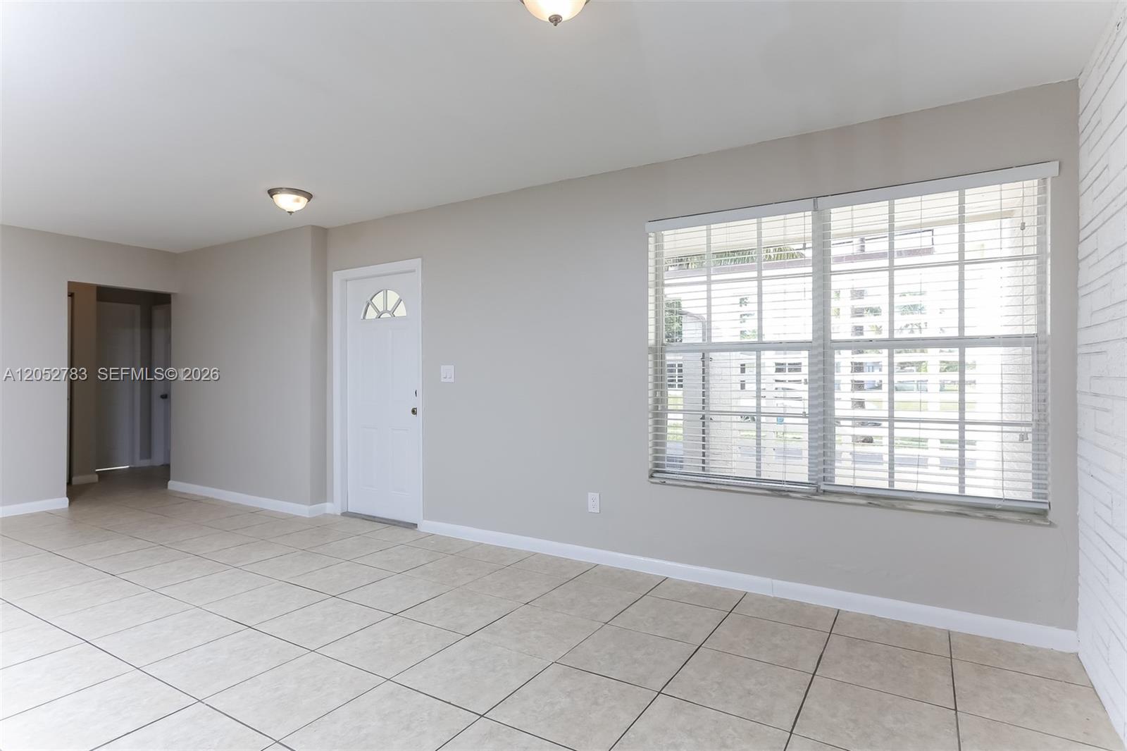 An empty room features light-colored tile flooring, beige walls, a white door, and large windows — 2230 NW 42nd Ave, Lauderhill FL 33313, LAUDERHILL, FL