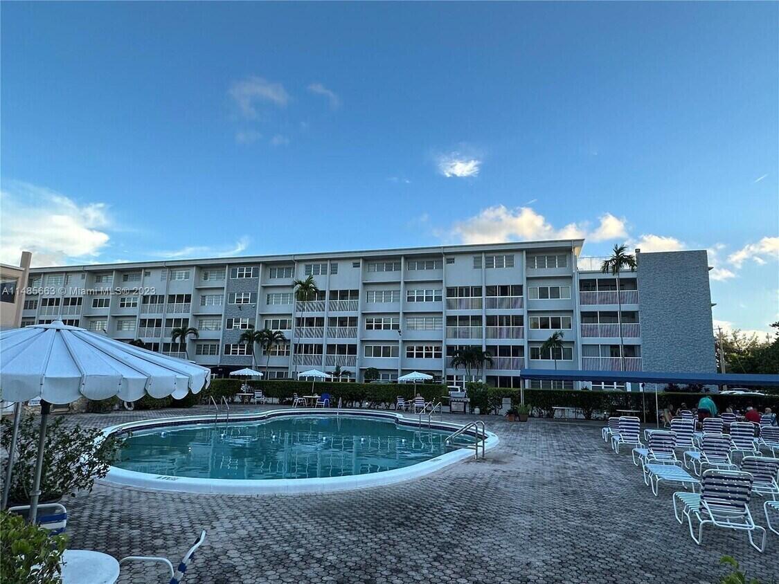 A swimming pool sits in front of a multi-story apartment building under a blue sky — 215 SE 3RD Avenue # 306D, Hallandale Beach FL 33009, HALLANDALE BEACH, FL