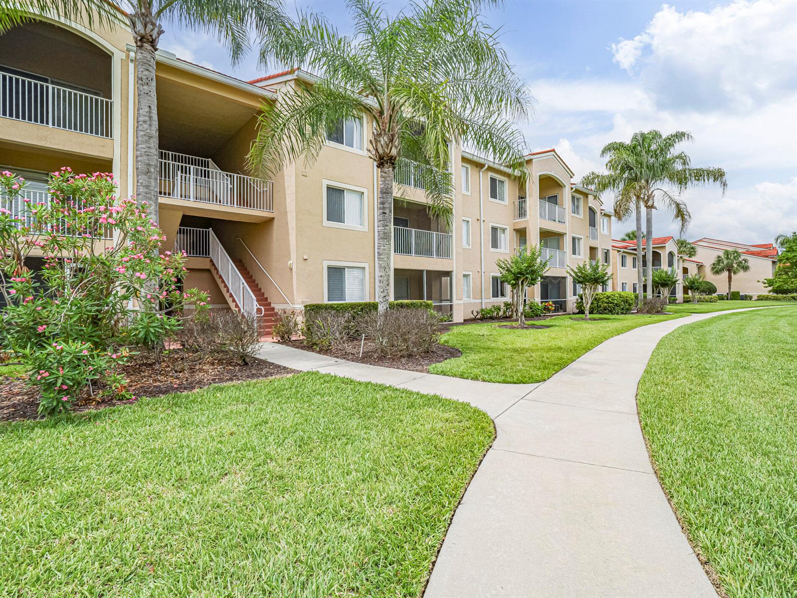 A multi-story apartment building with tan stucco walls, balconies, and palm trees along a sidewalk — VERO BEACH, FL