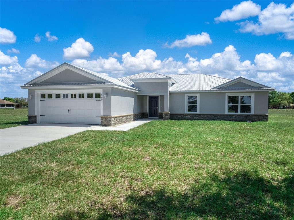 A single-story house with a white garage, gray stucco walls, stone accents, and metal roof — 7000 WOODMONT COURT, SEBRING, FL