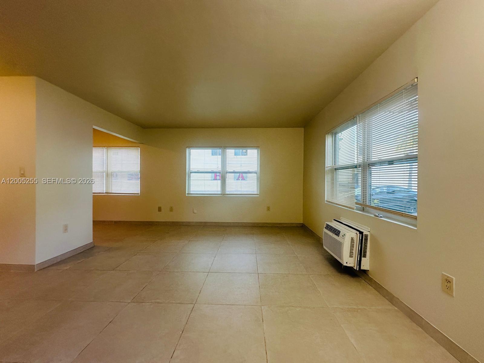An empty room features beige tile flooring, light-colored walls, multiple windows with blinds, and a wall-mounted air conditioner — 1708 Van Buren St # 6, Hollywood FL 33020, HOLLYWOOD, FL