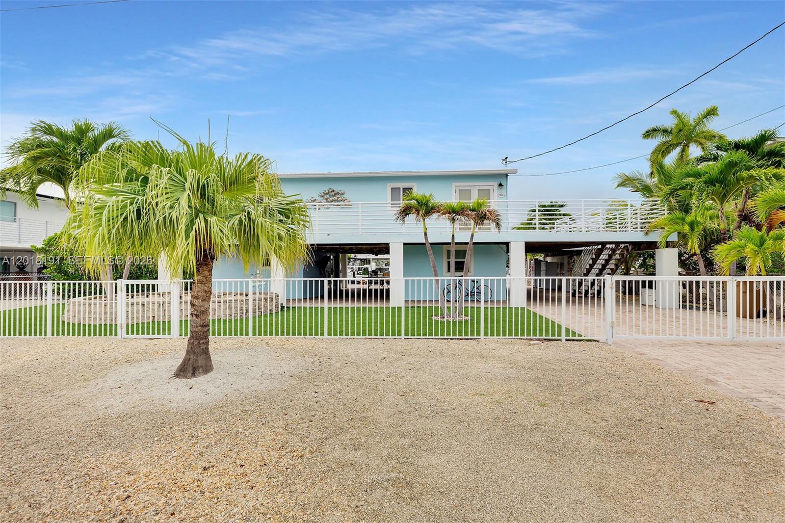 A light blue elevated house features a white fence, palm trees, and a gravel yard — 29565 Flying Cloud Ave, Big Pine FL 33043, BIG PINE, FL