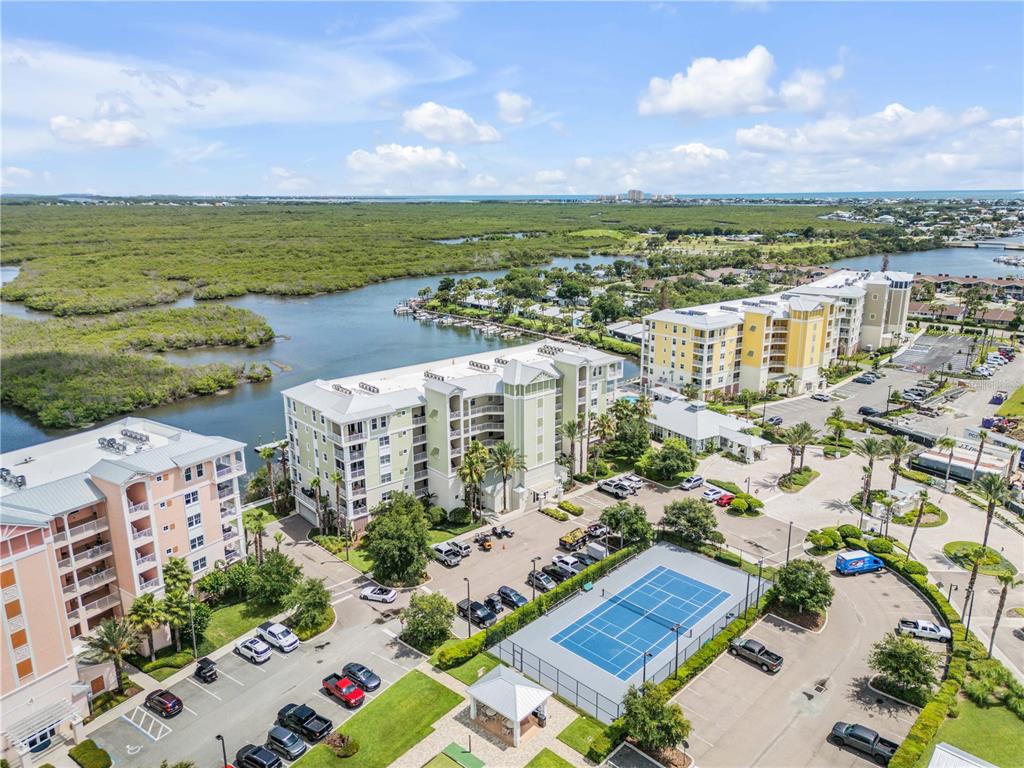 An aerial view shows multi-story apartment buildings, a tennis court, and a parking lot near water — 5 RIVERWALK DRIVE UNIT 506, NEW SMYRNA BEACH, FL