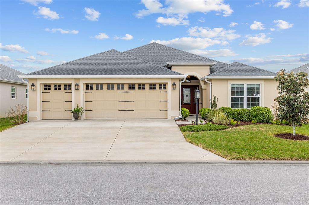 A beige single-story house features a three-car garage and a concrete driveway under a sky — 5791 HENRY LOOP, THE VILLAGES, FL