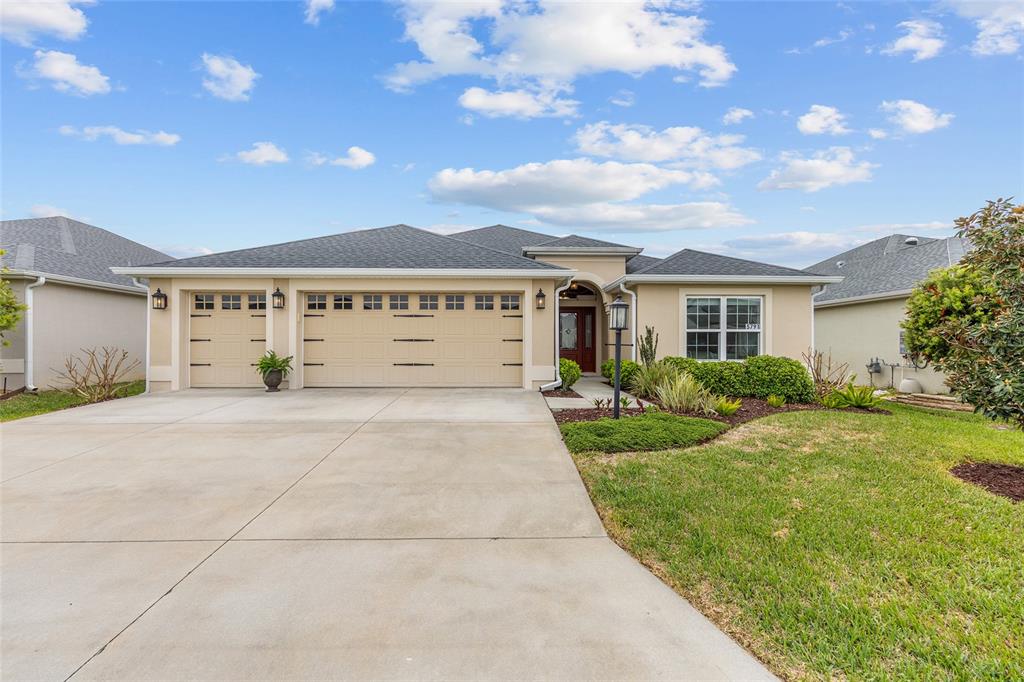 A single-story suburban house with a beige stucco exterior, two-car garage, and landscaped front yard — 5791 HENRY LOOP, THE VILLAGES, FL
