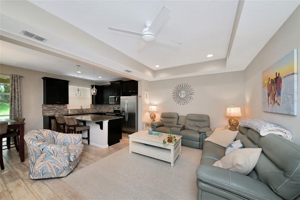 A living room with a tray ceiling and ceiling fan opens to a kitchen with dark cabinetry — 12455 TRAILHEAD DRIVE, BRADENTON, FL