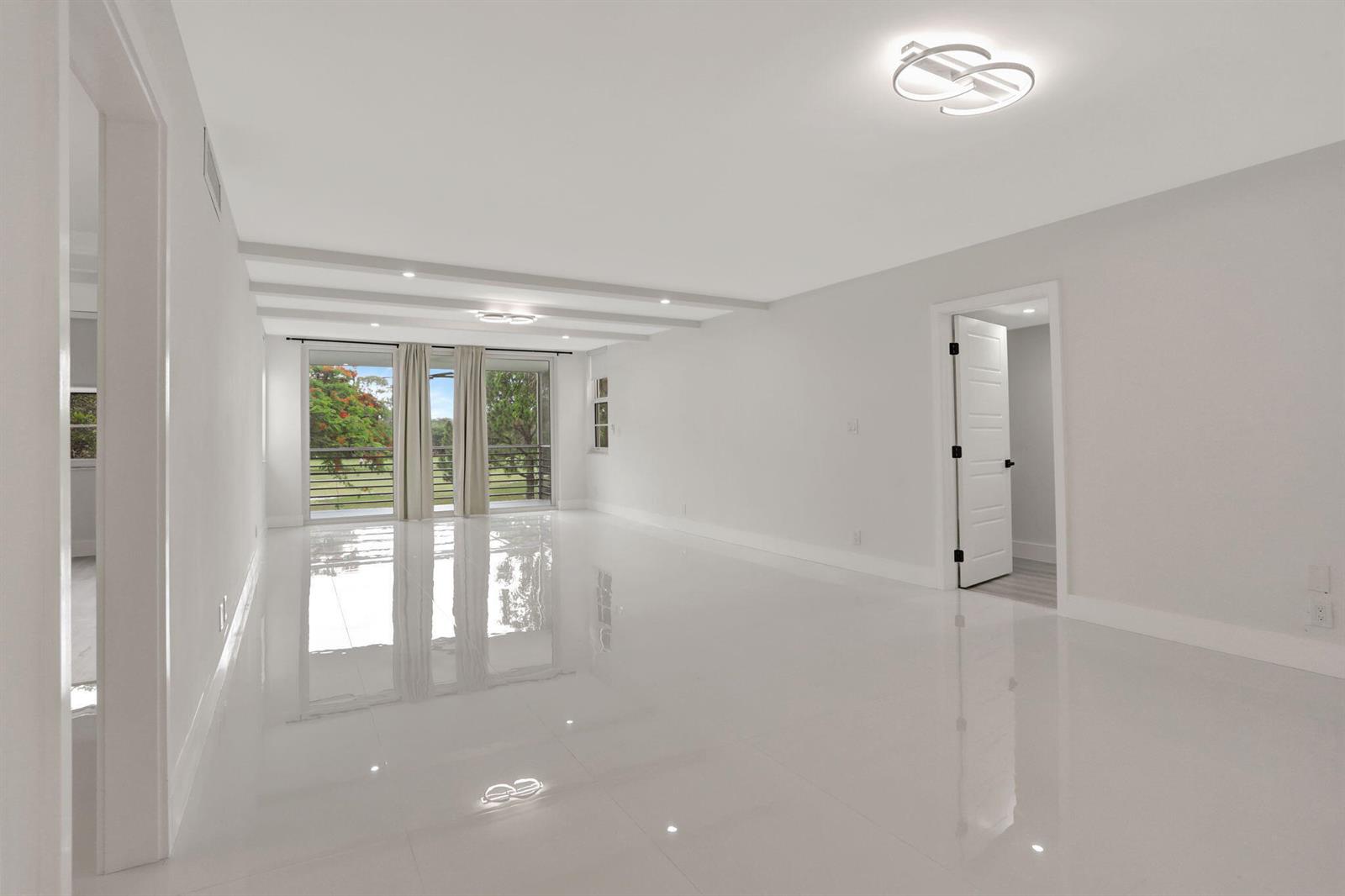 A bright, empty room features white tile floors, recessed lighting, and a balcony with greenery — 3505 Oaks Way # 204, Pompano Beach FL 33069, POMPANO BEACH, FL