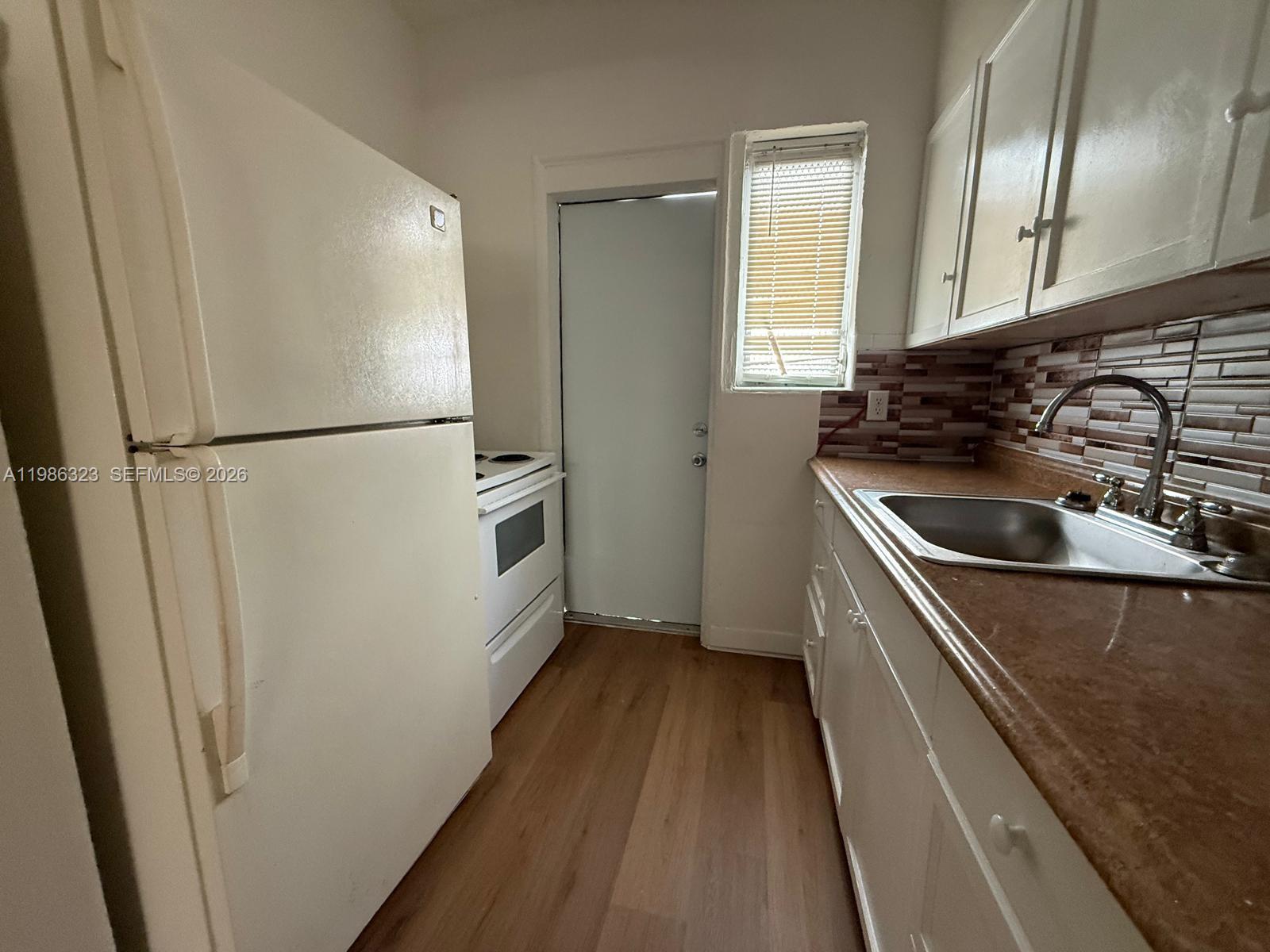 A small kitchen features white appliances, wood-look flooring, and a tiled backsplash — 6830 Harding Ave # 1, Miami Beach FL 33141, MIAMI BEACH, FL