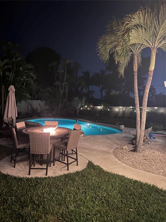 A backyard patio at night features a fire pit dining set, swimming pool, and palm trees — 6304 BEECHWOOD AVENUE, SARASOTA, FL
