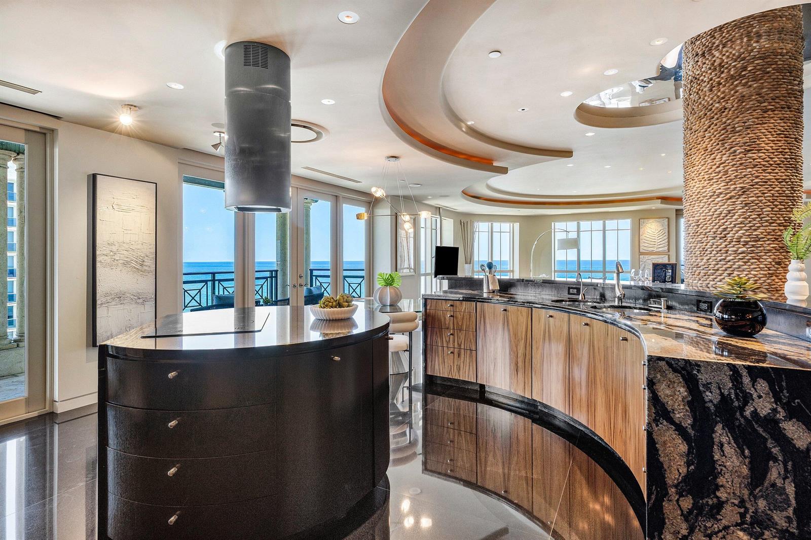 A modern kitchen features a curved island, wood cabinetry, stone countertops, and ocean views — 400 S Ocean Boulevard, Boca Raton FL 33432, BOCA RATON, FL