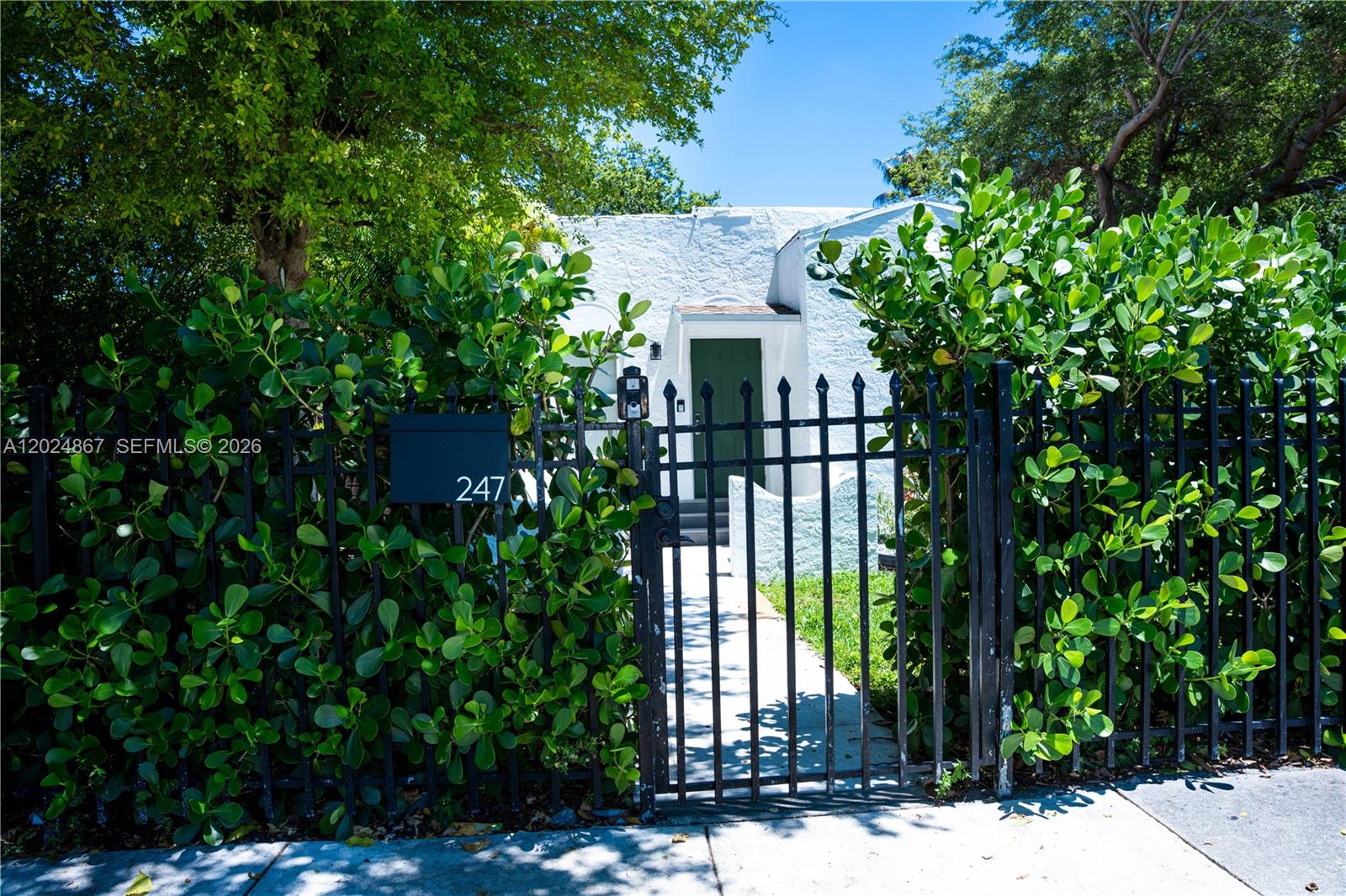 A white stucco house is partially obscured by lush green hedges behind a black fence — 247 NW 58th St, Miami FL 33127, MIAMI, FL