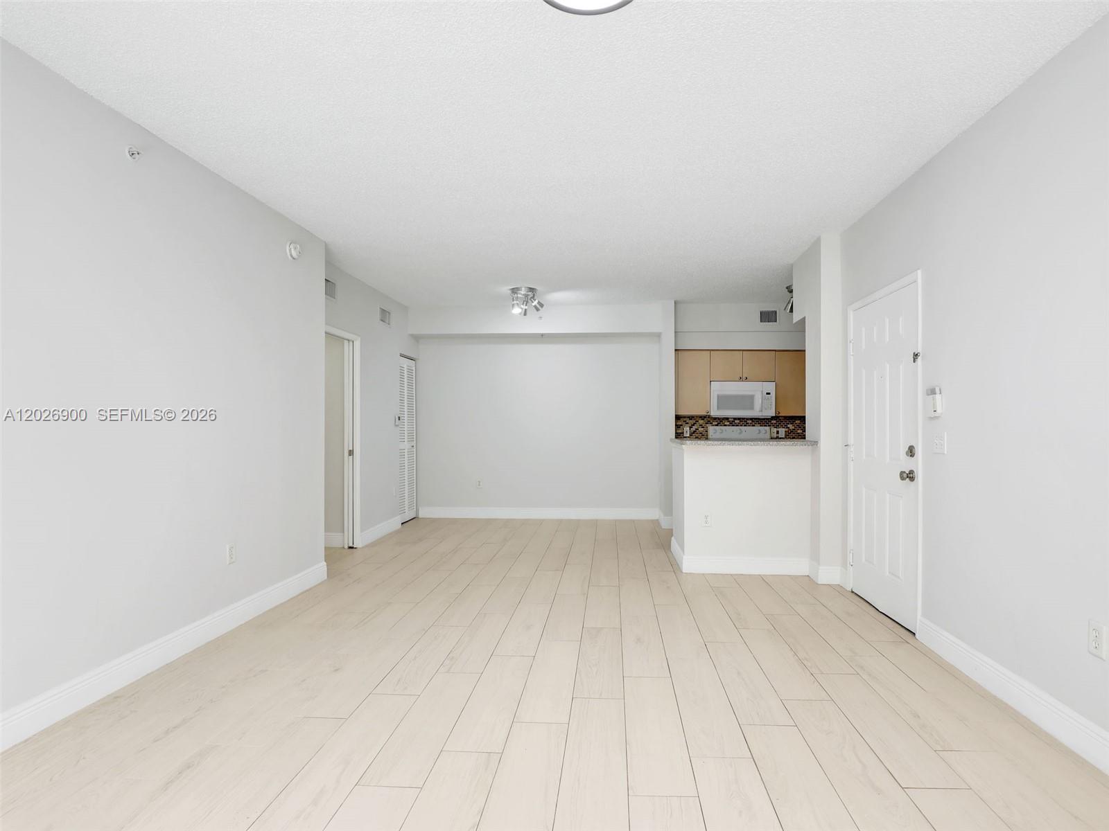 An open living area features light wood-look tile flooring, white walls, and an adjacent kitchen — 1235 Reserve Way # 107, Naples FL 34105, NAPLES, FL