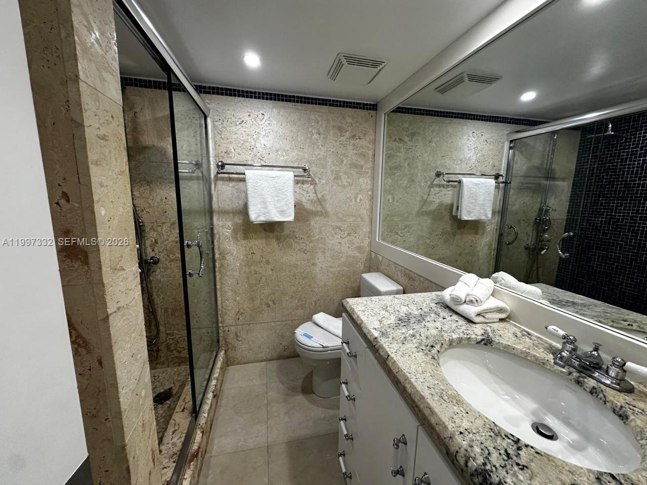 A bathroom features a glass-enclosed shower, a granite vanity, and light-colored stone wall tiles — 5225 Collins Ave # 614, Miami Beach FL 33140, MIAMI BEACH, FL