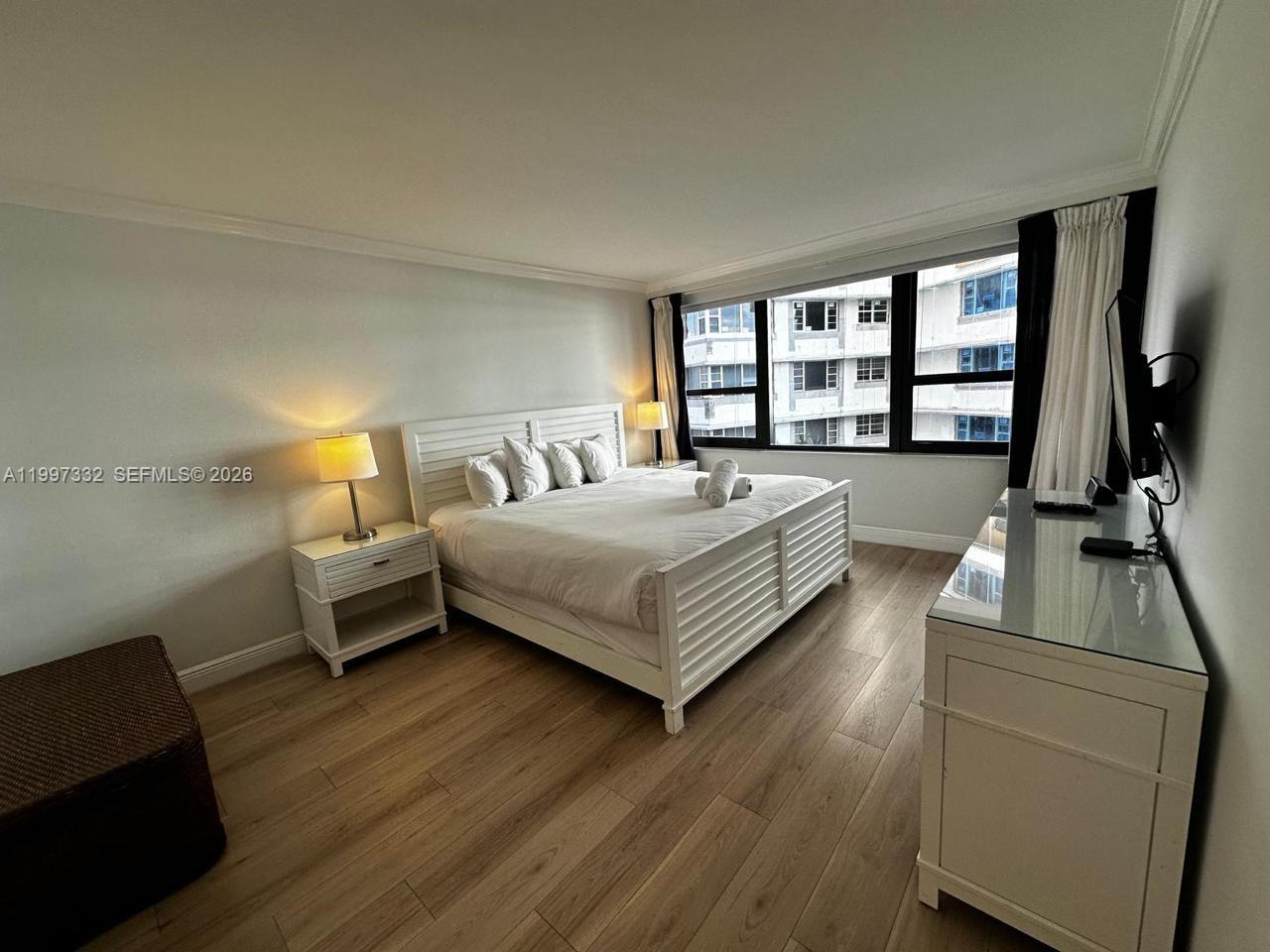 A bedroom features a white bed, matching nightstands, a glass-topped dresser, and large windows — 5225 Collins Ave # 614, Miami Beach FL 33140, MIAMI BEACH, FL