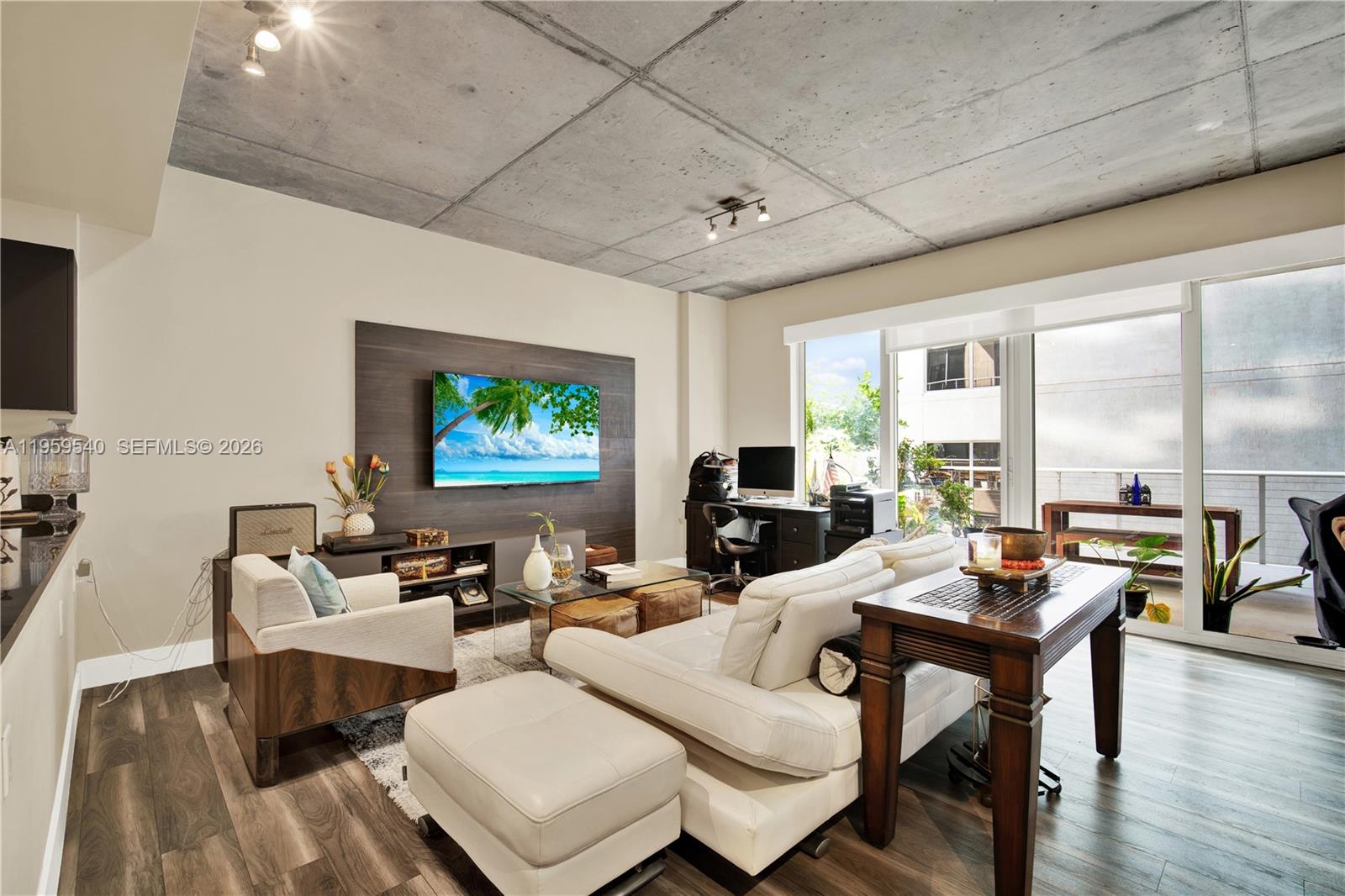 A modern living room features a concrete ceiling, wood flooring, and a balcony view — 3449 NE 1st Ave # 110, Miami FL 33137, MIAMI, FL