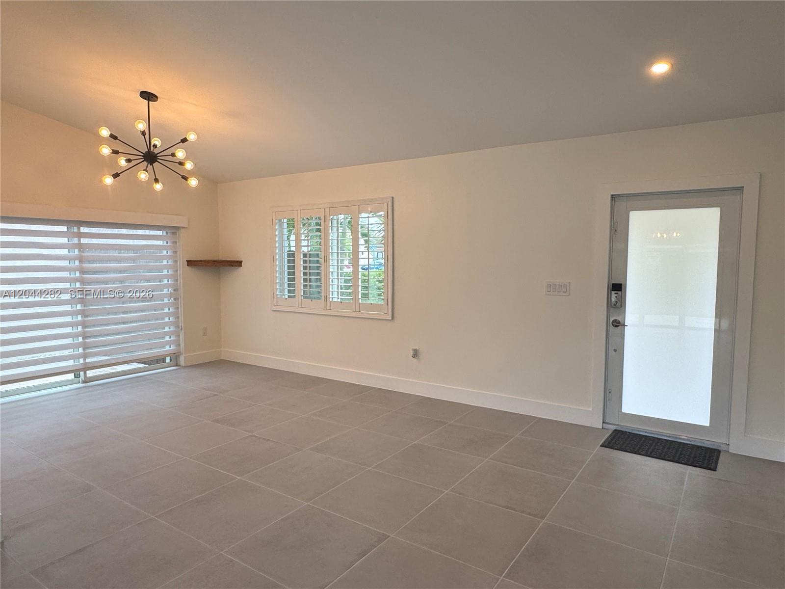 A bright, empty living room features large gray floor tiles, white walls, and a chandelier — 12845 SW 150th Ter, Miami FL 33186, MIAMI, FL