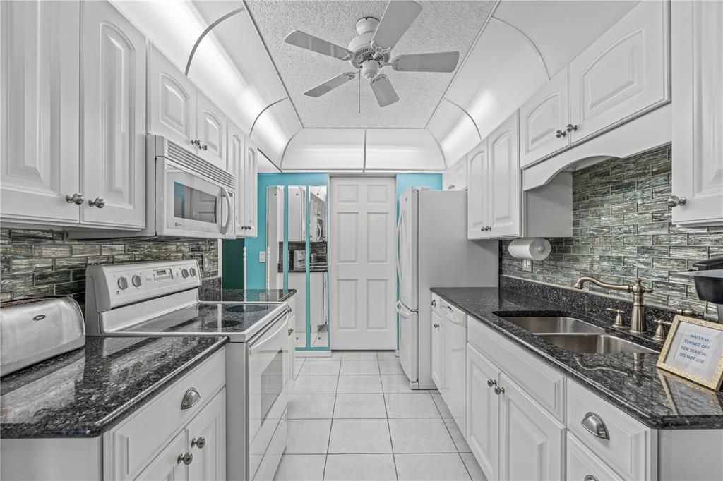 A galley kitchen features white cabinets, dark granite countertops, a tiled backsplash, and white appliances — 101 BENT TREE DRIVE UNIT 15, DAYTONA BEACH, FL