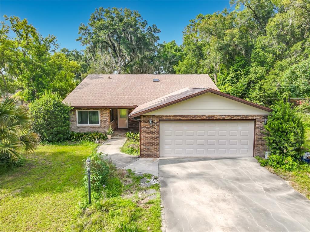 A single-story brick house with a two-car garage, concrete driveway, and surrounding mature trees — 474 W OAKWOOD AVENUE, ORANGE CITY, FL