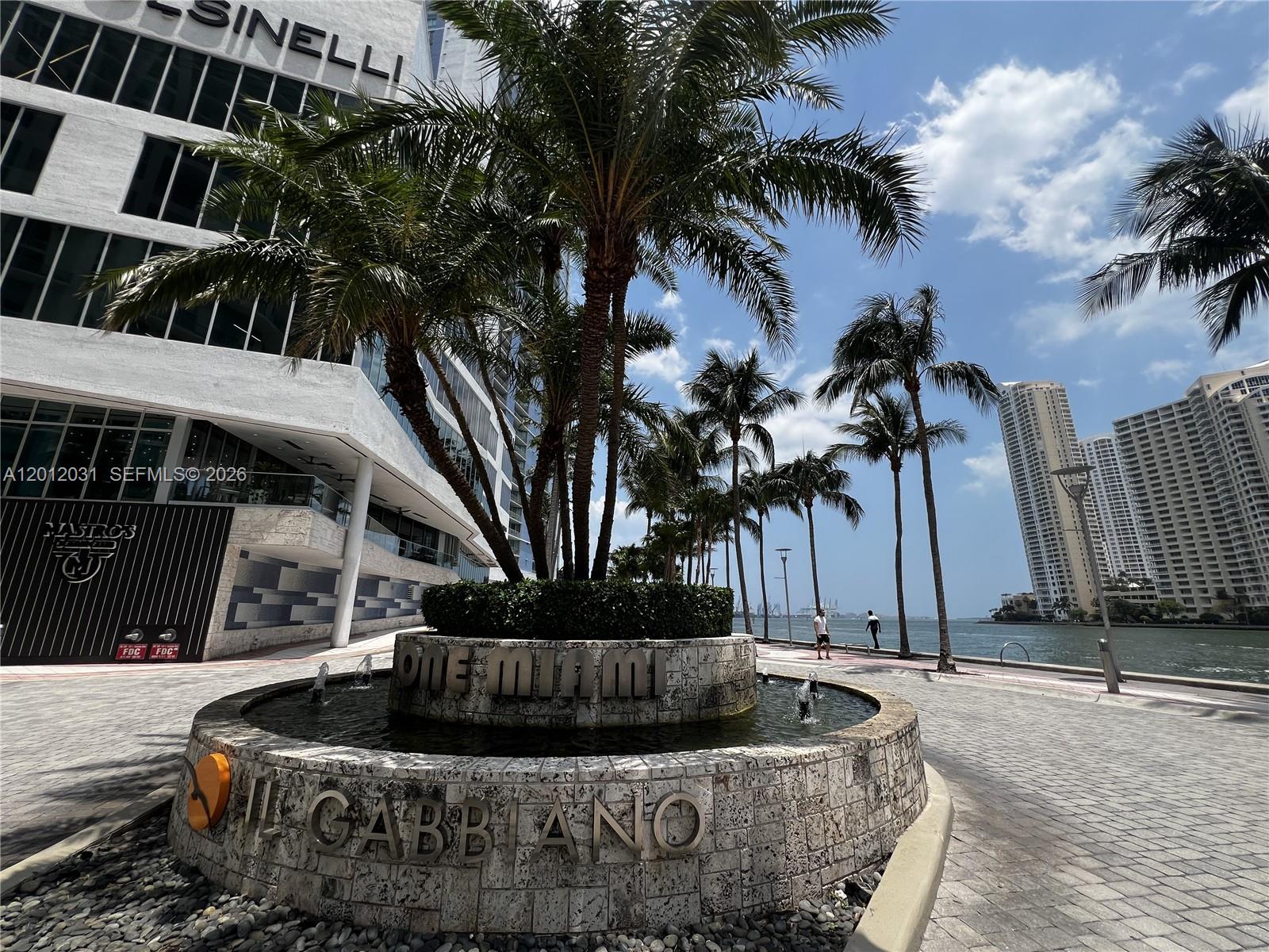A stone fountain with palm trees sits on a paved waterfront plaza near tall buildings — 335 S Biscayne Blvd # 2900, Miami FL 33131, MIAMI, FL