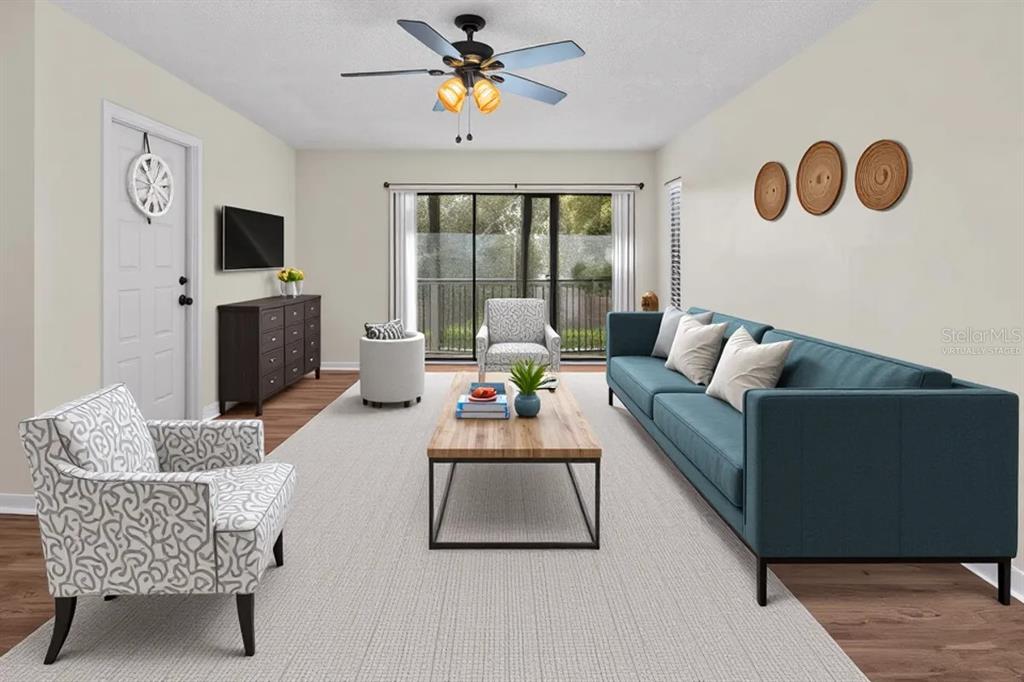 This virtually staged living room features a blue sofa, patterned armchairs, and a sliding glass door — 1127 PINE RIDGE CIRCLE W UNIT A2, TARPON SPRINGS, FL