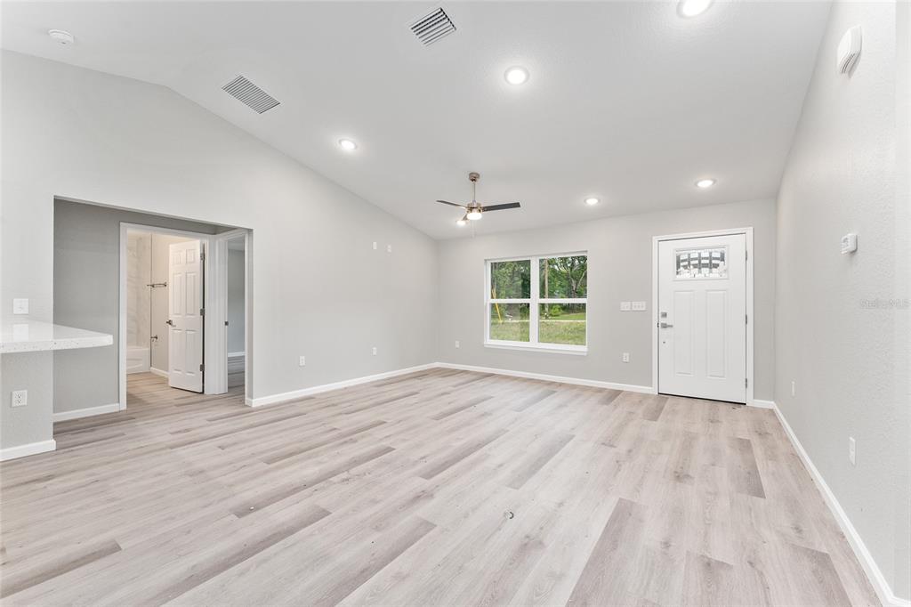 A bright, empty living room features light-colored wood-look flooring, a vaulted ceiling, and a front door — 10374 SE 126TH LANE, BELLEVIEW, FL